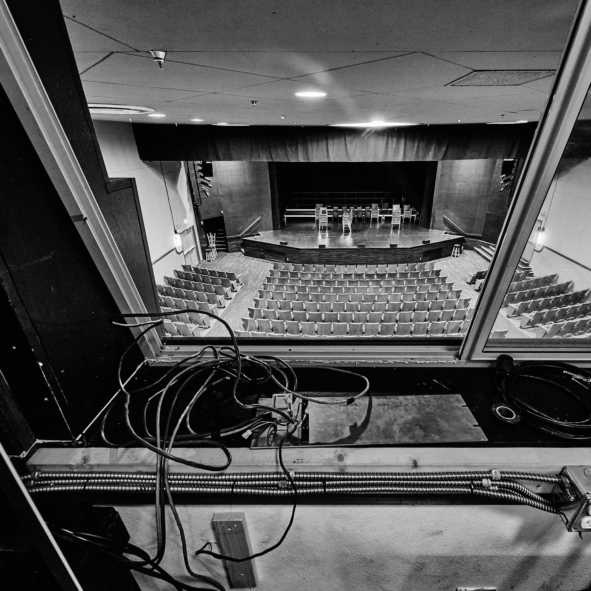 Black and white photo of an empty stage with stacked chairs and scattered equipment, viewed from a balcony or balcony-like structure.