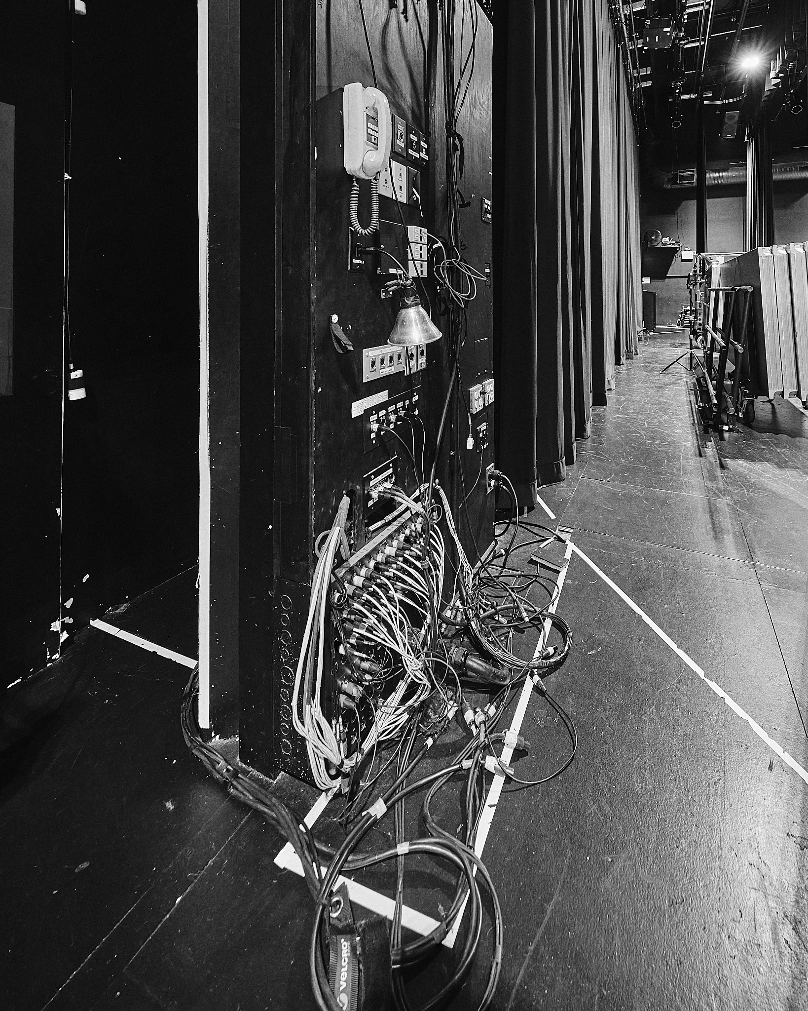 A stage scene with a black backdrop, featuring a complex setup of cables, switches, and outlets attached to a black panel, with a white telephone and various electronic devices. Some cables are draped onto the stage floor, with equipment carts visibl