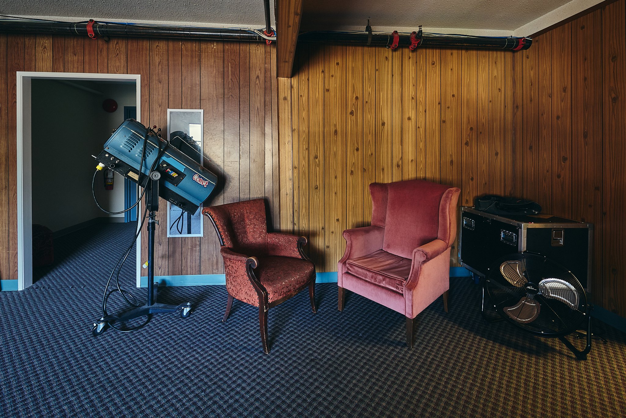 A room with wooden paneled walls and a blue and black patterned carpet. There are vintage-style chairs with red and pink upholstery, a black case, a fan, and a lighting setup with a blue Comet light on a stand.