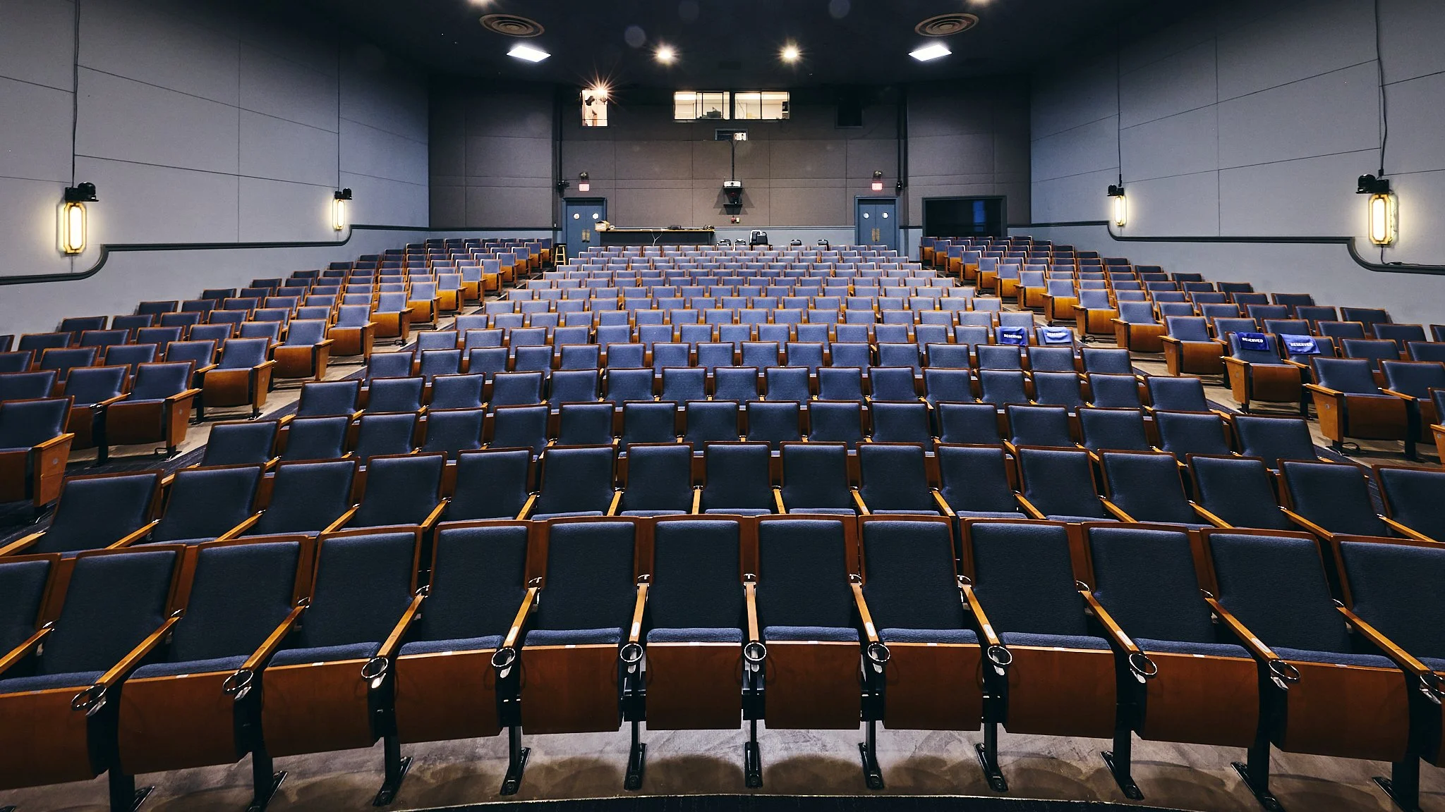 Empty auditorium with rows of blue and wooden chairs, gray walls, and black ceiling lights.