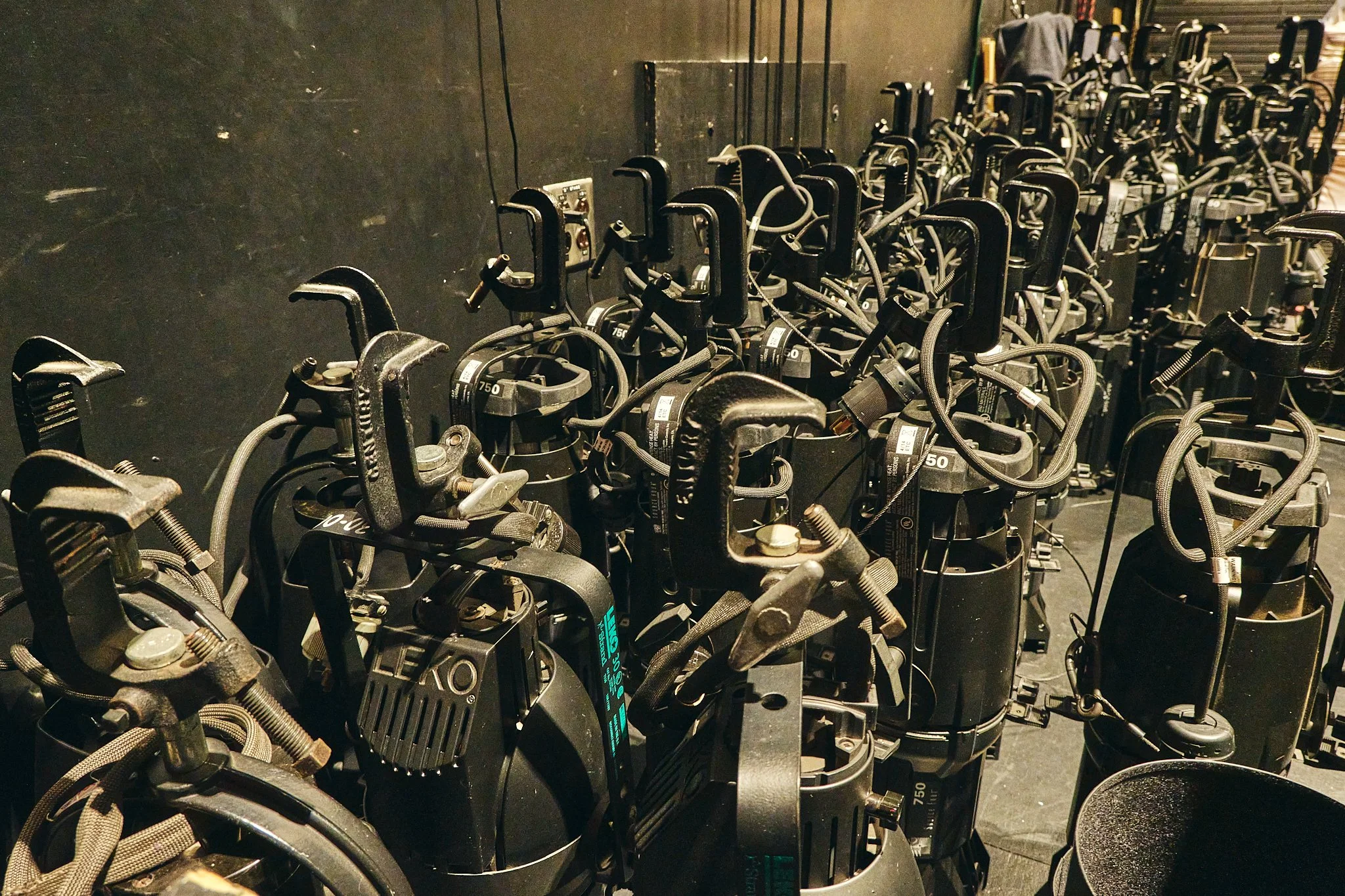 Row of black power tools with cords, stacked in a storage area with a dark wall in the background.