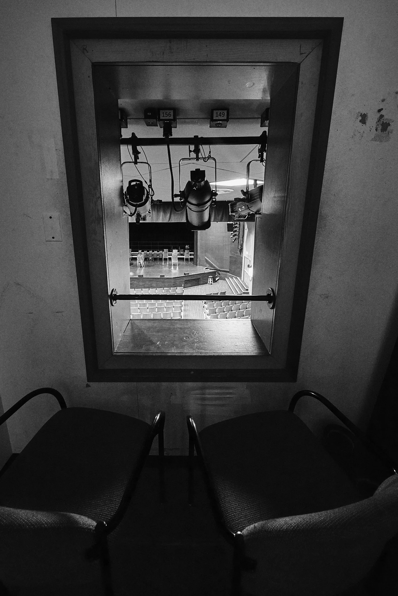 Black and white view downward through a fire alarm pull station covering, showing a stage and empty theater seats.