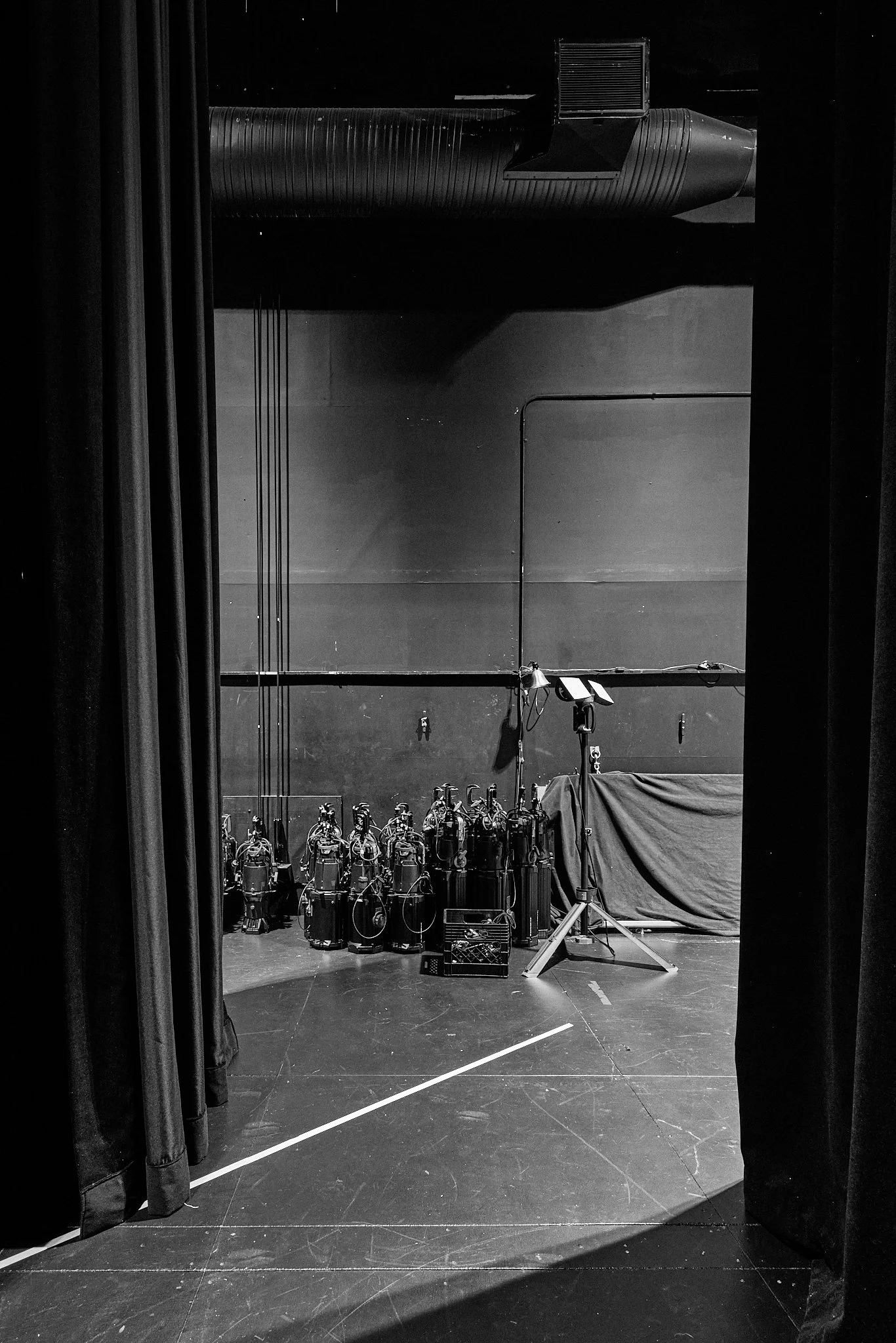 Black and white photo of a stage with equipment including numerous stage lights, a covered object, and a wall with hanging cables, viewed from behind stage curtains.