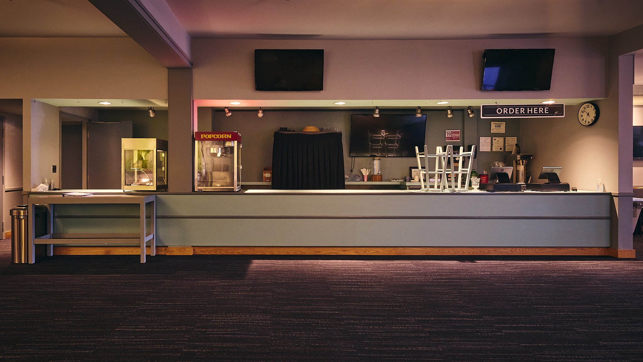 Empty concession or snack counter at a movie theater with popcorn machine, signs, and television screens hanging above.