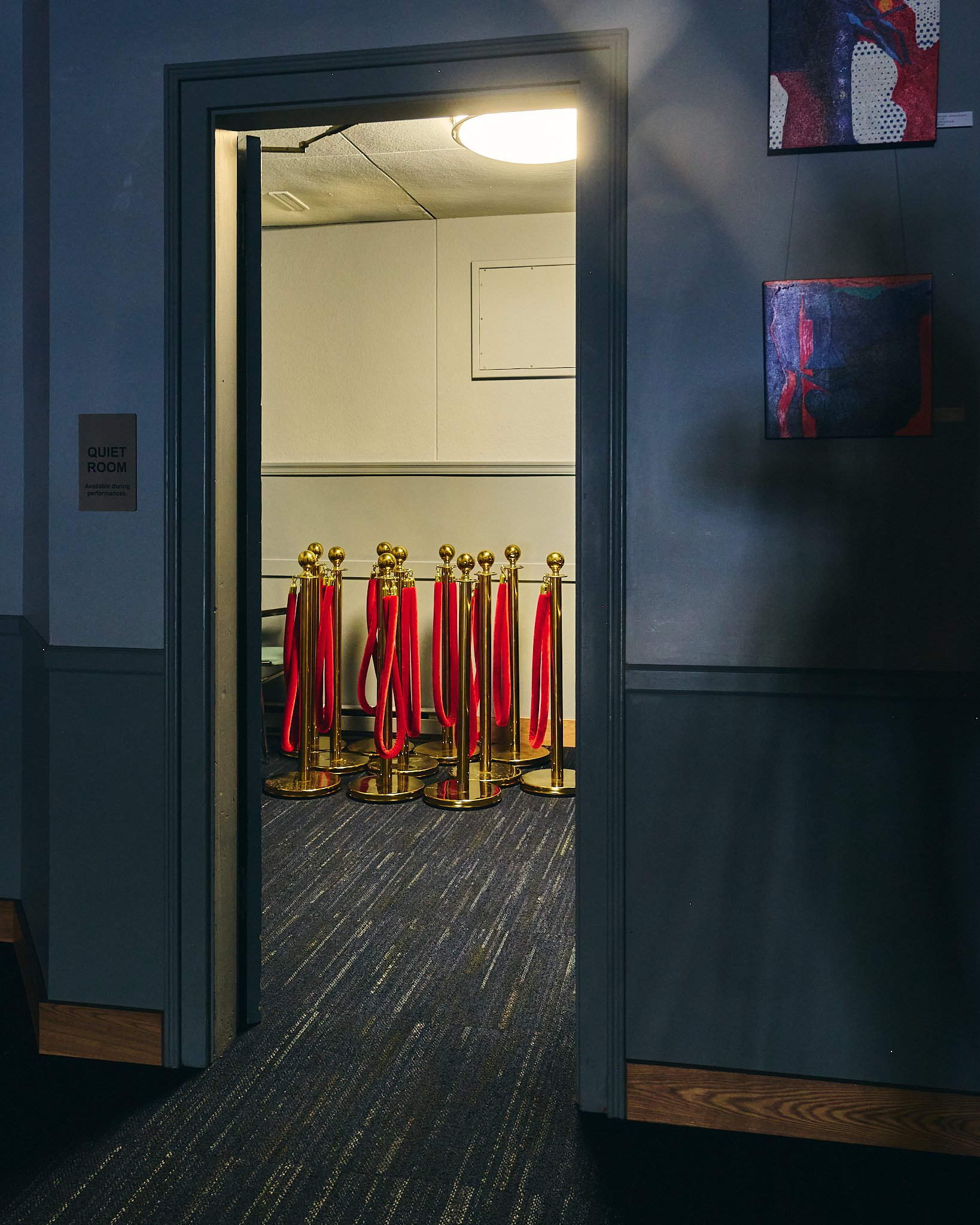 View of an art gallery with paintings on the wall, showing the open door of a room with yellow posts and red velvet ropes blocking entrance, a sign stating "Quiet Room," and a section of dark-colored carpeted flooring.