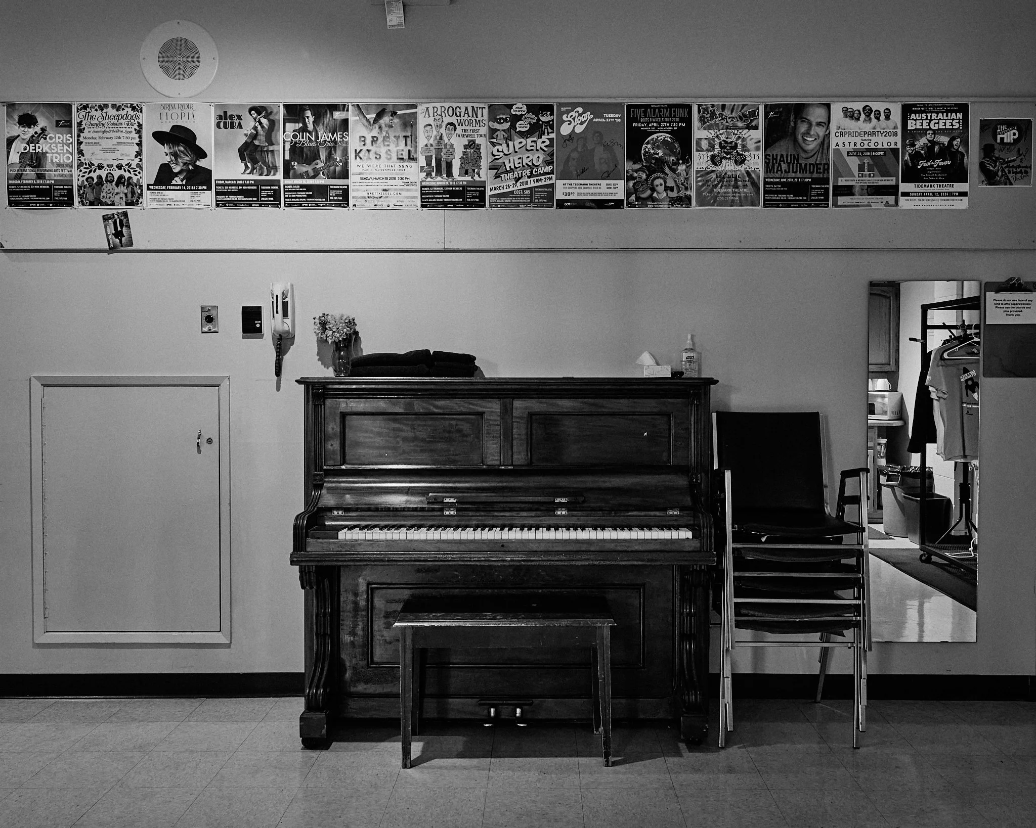 Black and white photo of a wall with a row of promotional posters for music events and performances, an open doorway showing a part of a room with clothing on racks, and a piano with some items on top, such as a vase of flowers, a cloth, a sanitizer 