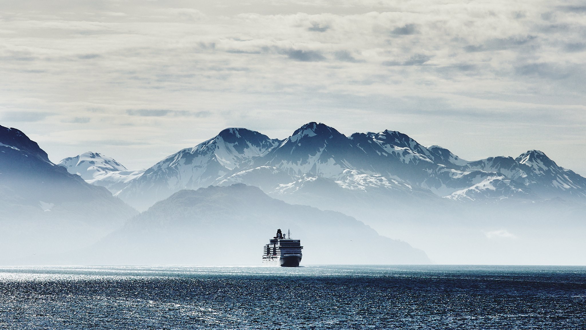 A cruise ship sailing in the ocean with snow-capped mountains and cloudy sky in the background.