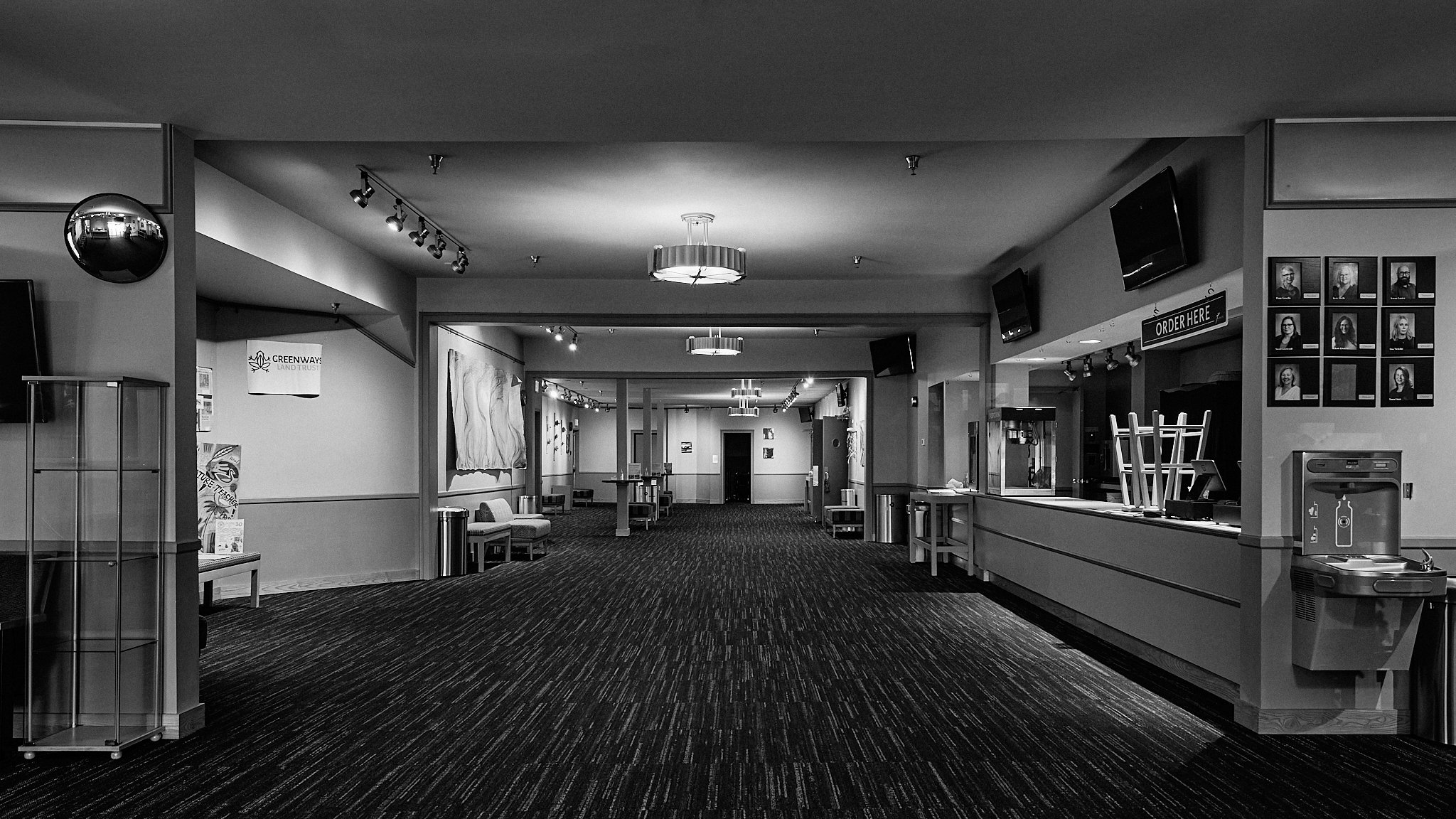 Empty lobby or waiting area of a theater or entertainment venue with chairs, artwork on the walls, a sign that says 'Order Here,' and a counter with a few items on it.