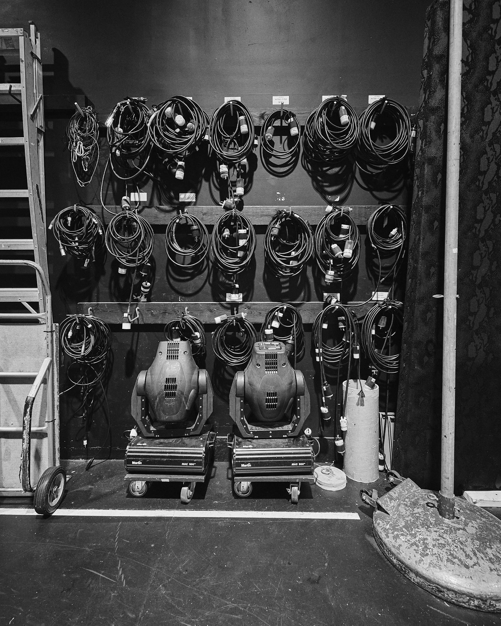 Black and white photo of a wall with multiple cables hanging and coiled, two industrial machines at the bottom, a ladder on the left, and equipment on the right in a workshop or stage setting.