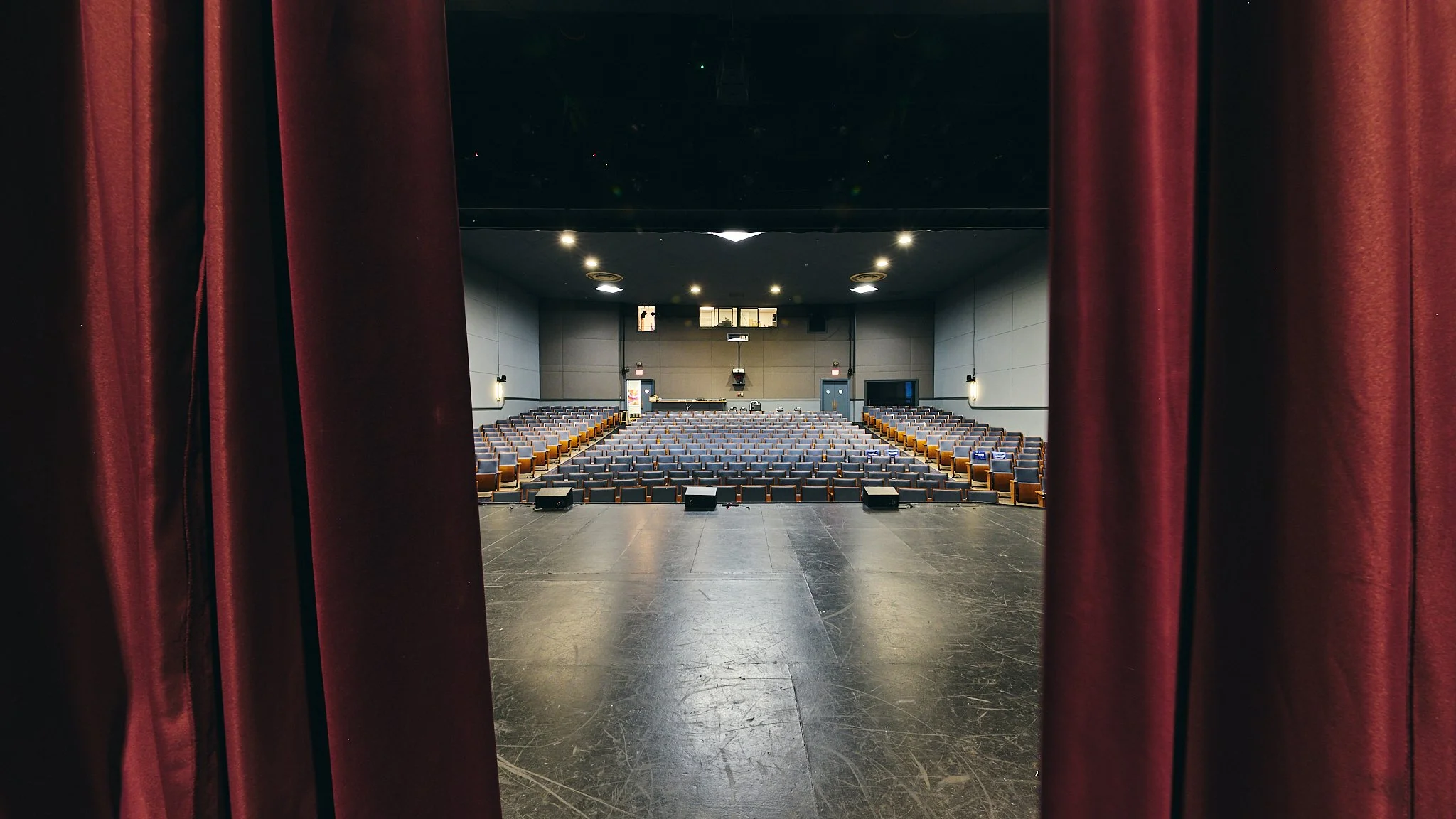 View of an empty theater auditorium through stage curtains, showing rows of seats facing a screen and stage area.