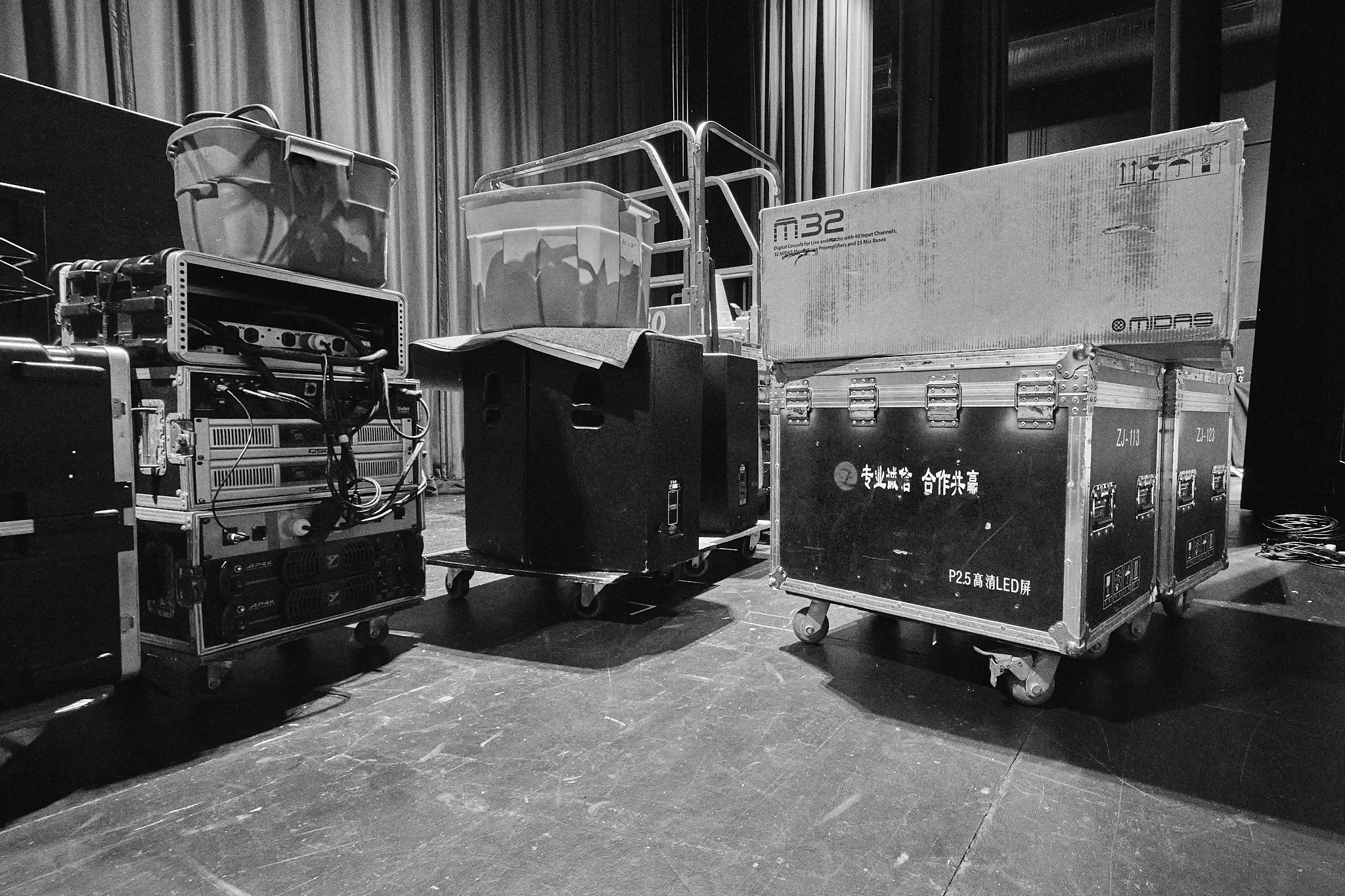Black and white photo of equipment cases, a cart with plastic storage bins, and boxes on stage or backstage.