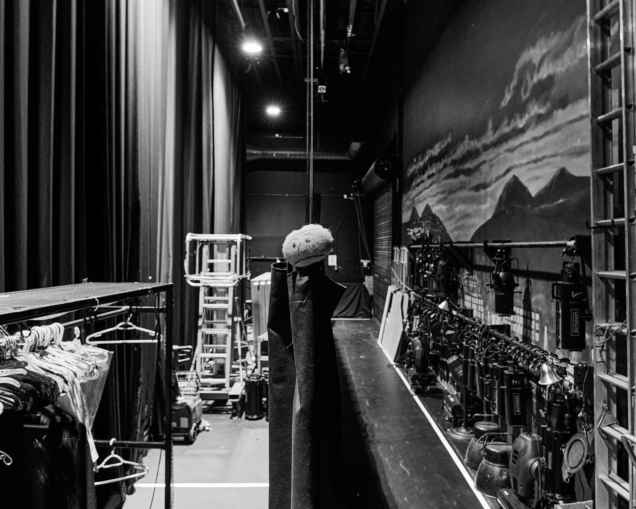 Black and white photo of a theater stage with set pieces including mountain backgrounds and stage lighting, taken from backstage.