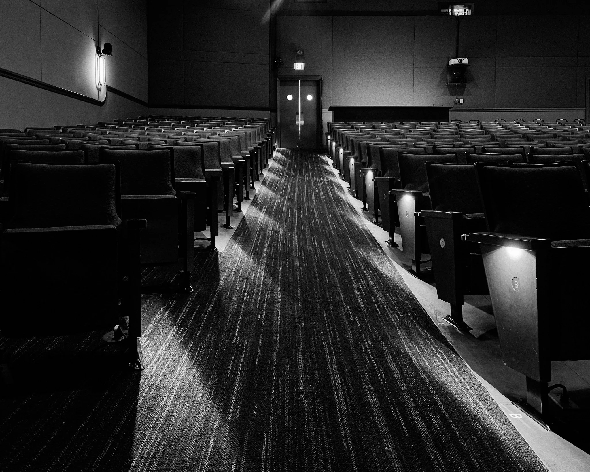 Empty movie theater with rows of chairs, a dark carpet, walls, and a double exit door at the back with illuminated exit sign and lights, in black and white.