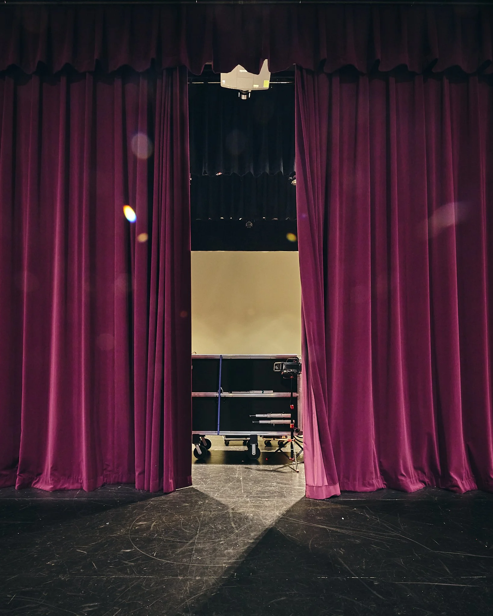Stage viewed through purple theater curtains, with part of the stage visible, including a wheeled platform, lighting equipment, and a black backdrop.