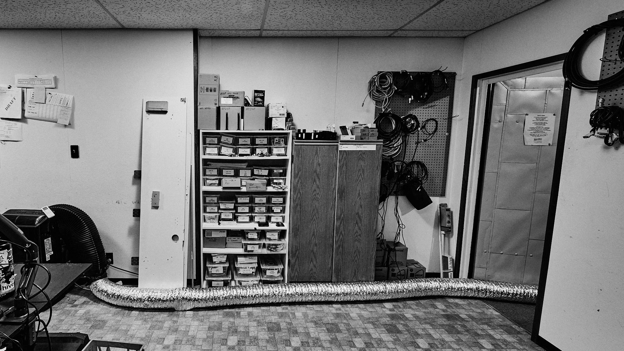 A black and white photo of a room with various electronic components, cables, and equipment organized on shelves and pegboards.