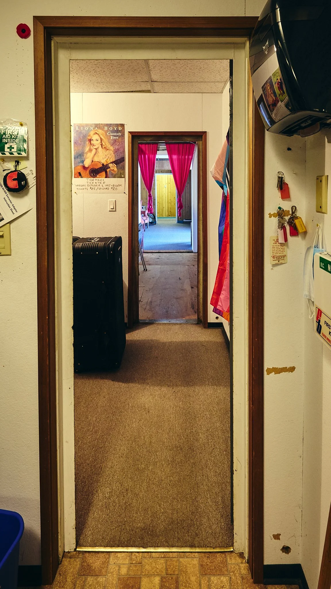 A doorway leading to a room with pink curtains and a wooden fence outside. The hallway has a tan carpet and is decorated with various items on the walls, including a poster of a woman with a guitar and several keys hanging on hooks.