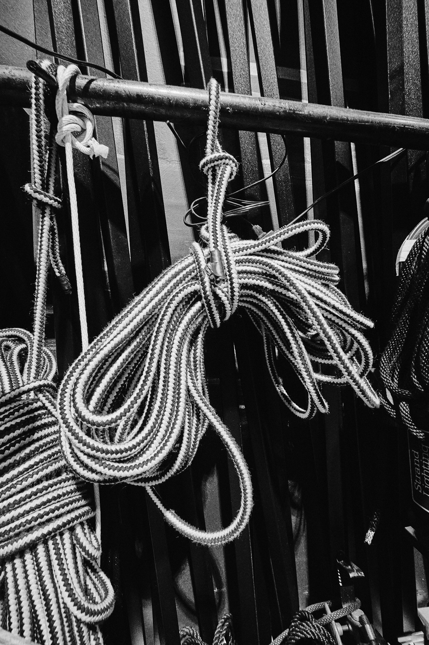 Black and white photo of several coiled and knotted ropes hanging on a metal railing, with a dark background.