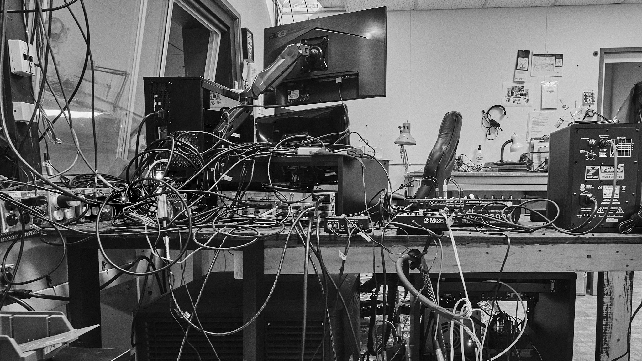 A cluttered workstation with multiple electronic devices, tangled cables, monitors, and audio equipment in a workshop or studio setting.