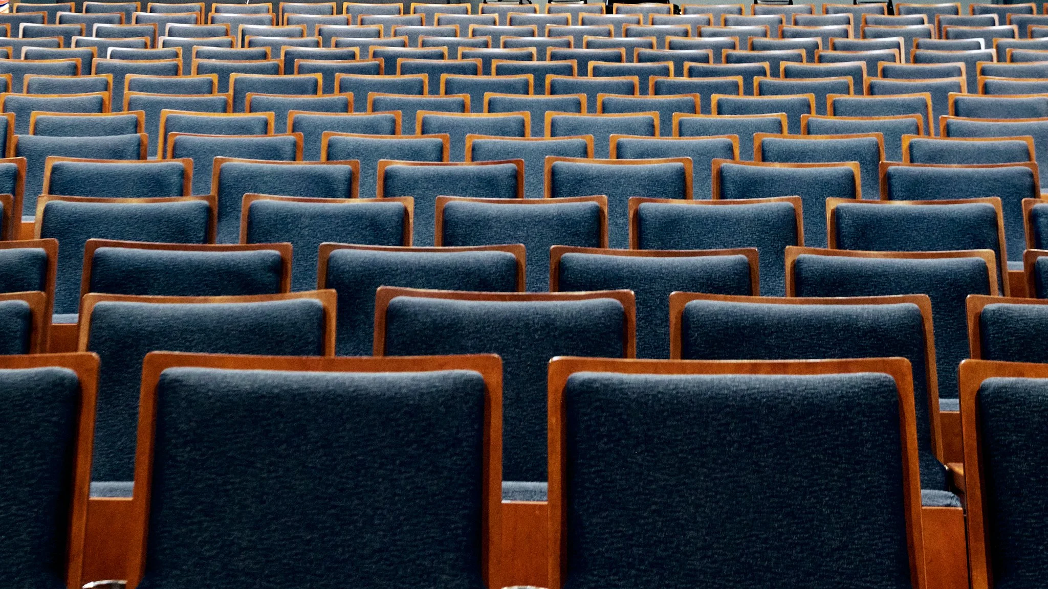 An empty auditorium filled with rows of blue upholstered chairs with wooden frames.