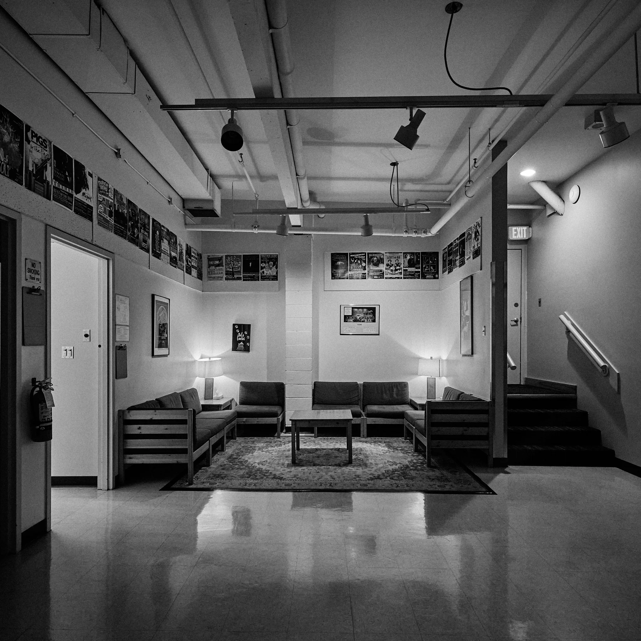 A black-and-white photo of an indoor waiting area with couches, tables, posters on the walls, and lighting fixtures on the ceiling.