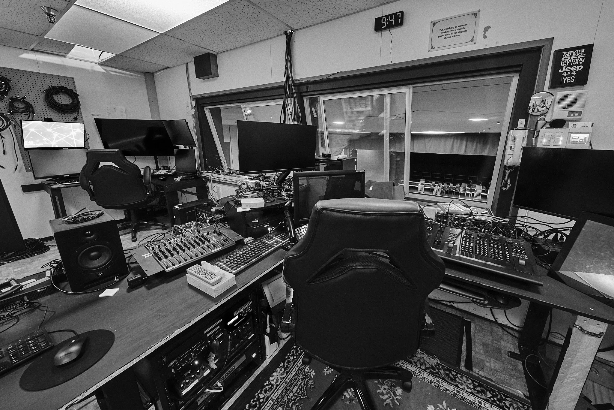 A cluttered sound recording studio with multiple monitors, audio equipment, wires, and a chair in front of a large window overlooking another room.