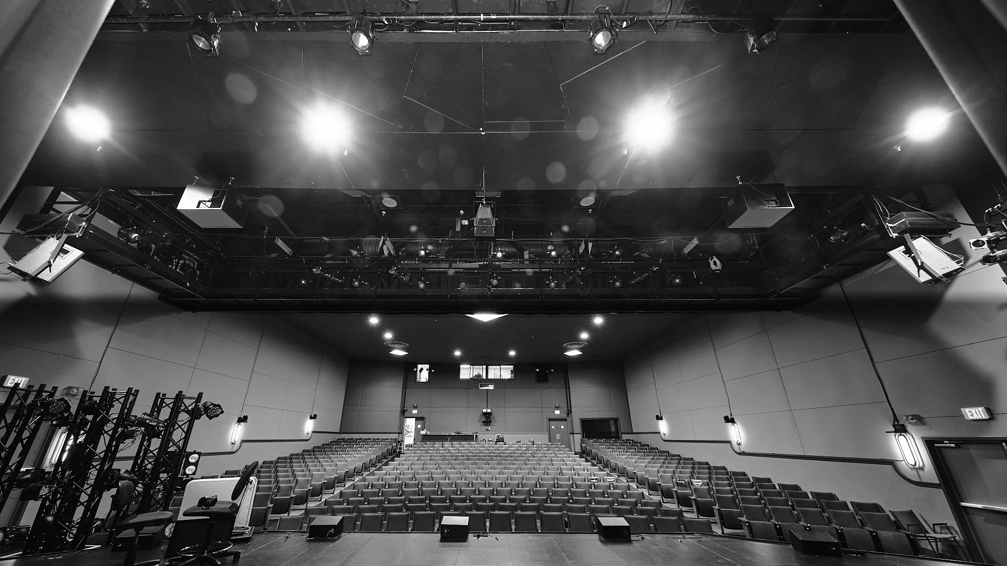 Empty theater or auditorium with rows of seats, stage with equipment, and overhead lighting and sound system.
