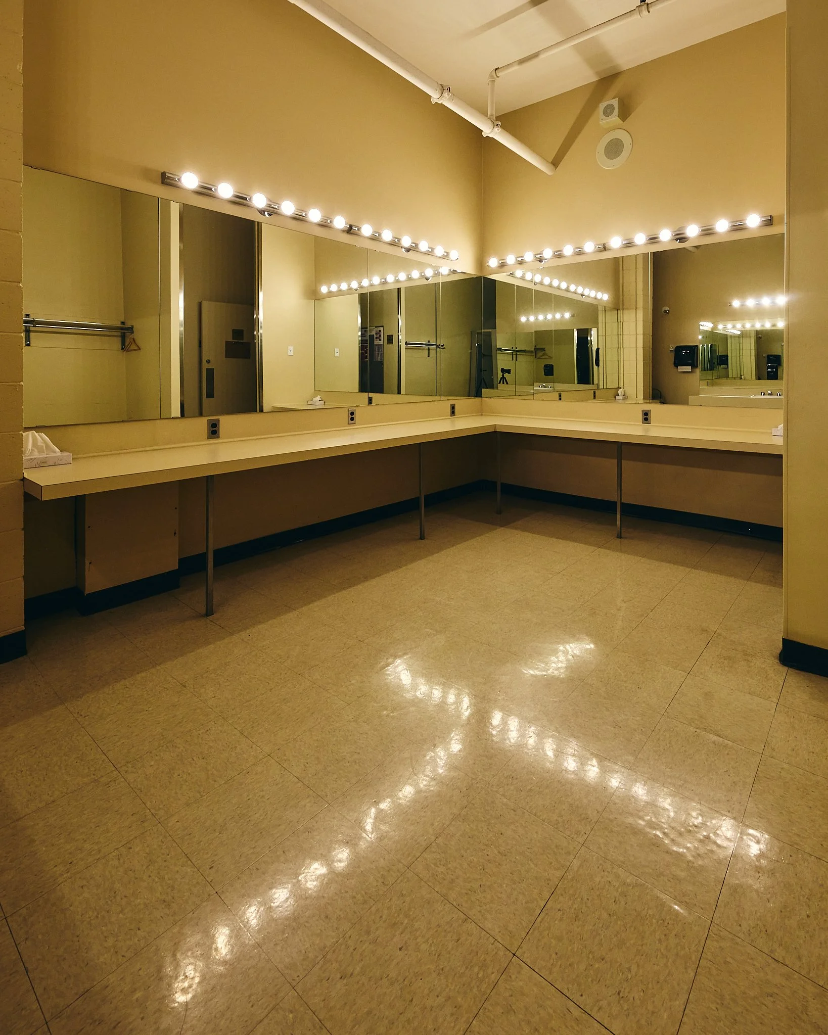 Empty dressing room with beige tiled floor, beige walls, large mirror with vanity lights, tissue box on counter, and a towel rack.