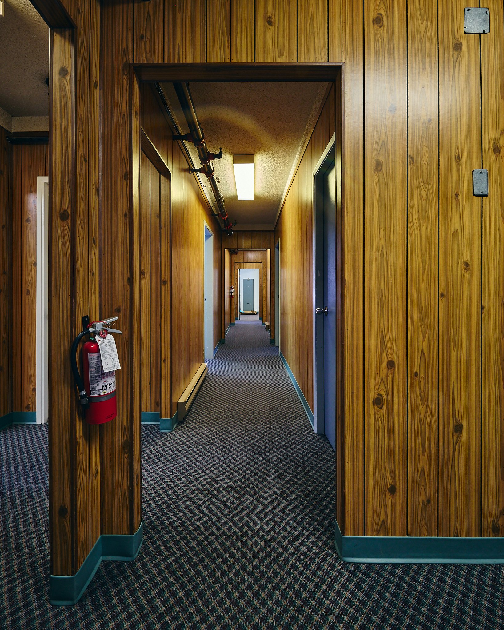 Wood-paneled hallway with multiple doors, carpeted floor, and fire extinguisher mounted on the wall on the left.