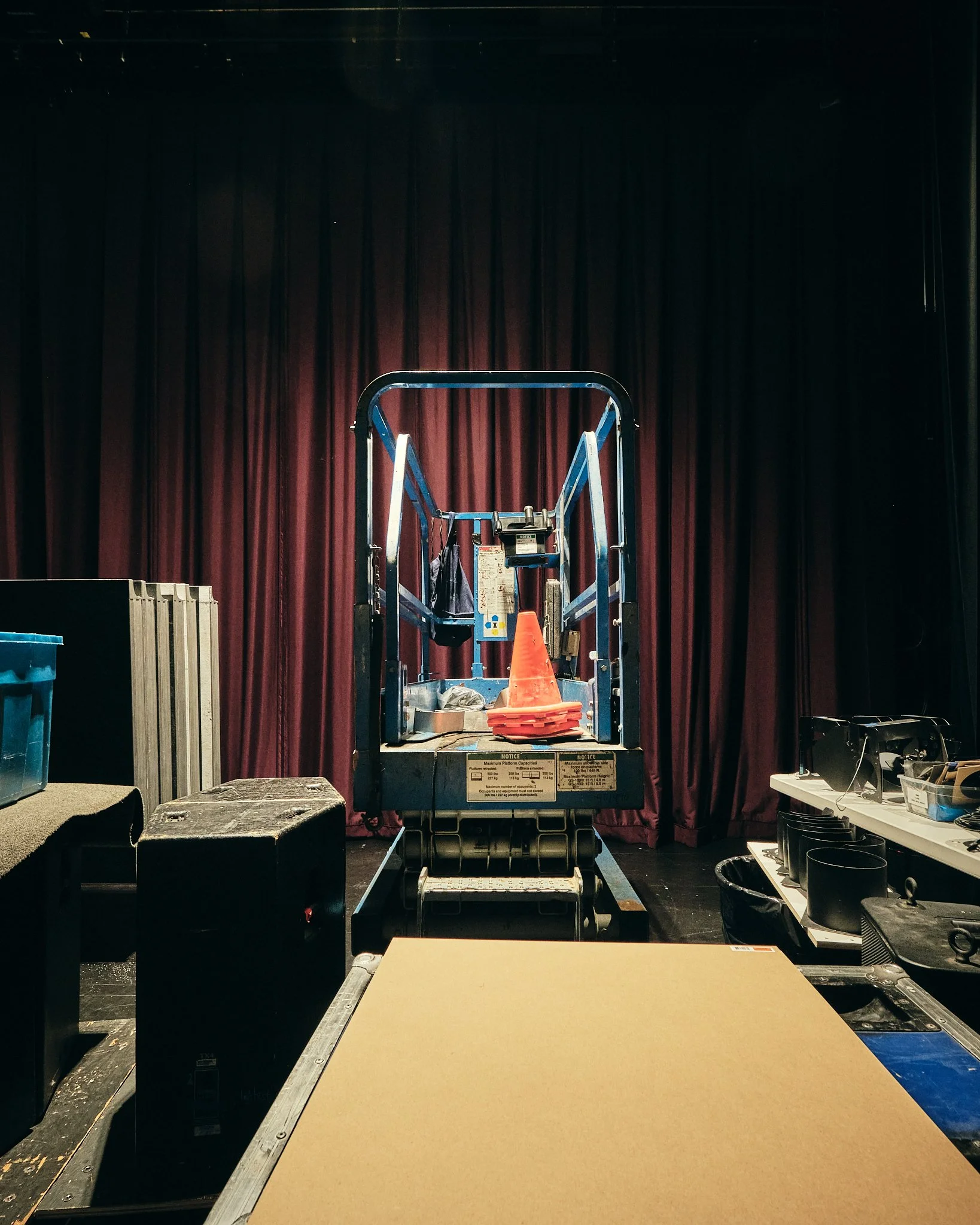 Stage with red curtains, stage lighting, a blue lift in the center with orange traffic cones on it, and various equipment and supplies surrounding the area.