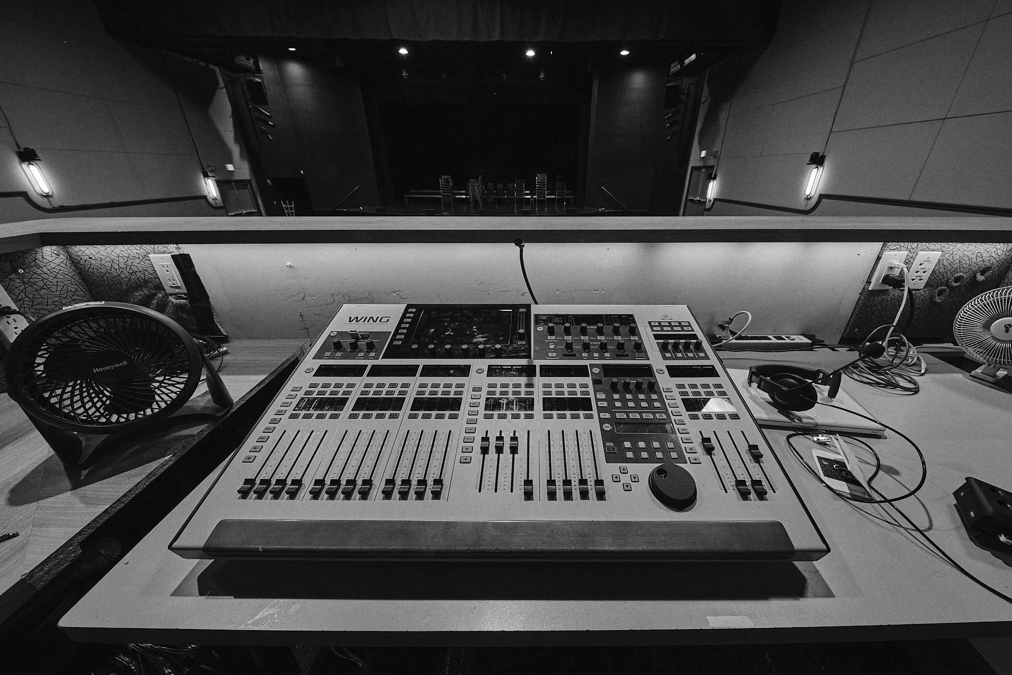 A DJ mixing console on a table with turntables, a fan, and audio equipment, in a dimly lit room with chairs visible in the background.