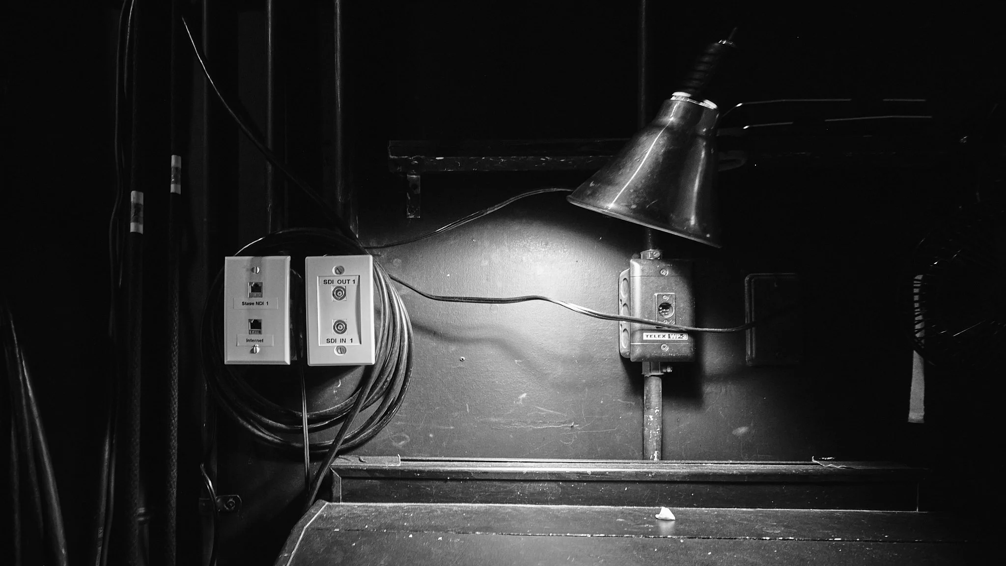 Black and white image showing a wall with electrical and cable boxes, and assorted wires, with a metallic lamp pointed downward.