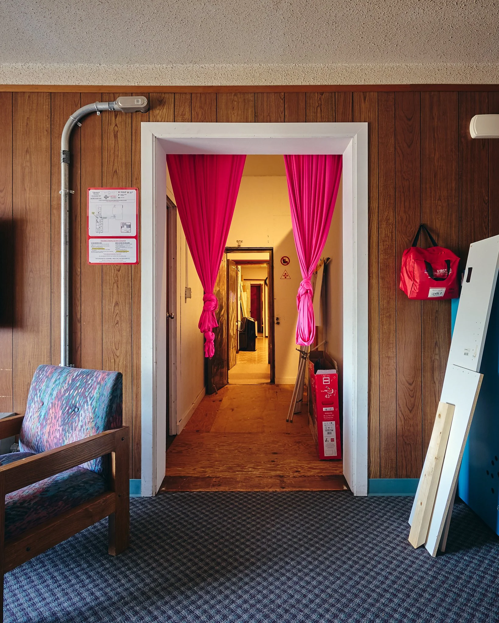 A wooden wall with a doorway draped with pink curtains leading to a hallway with a wooden floor. To the left, there is a cushioned wooden bench. To the right, a red bag hangs on the wall near a white frame. There is a fire extinguisher and a sign abo