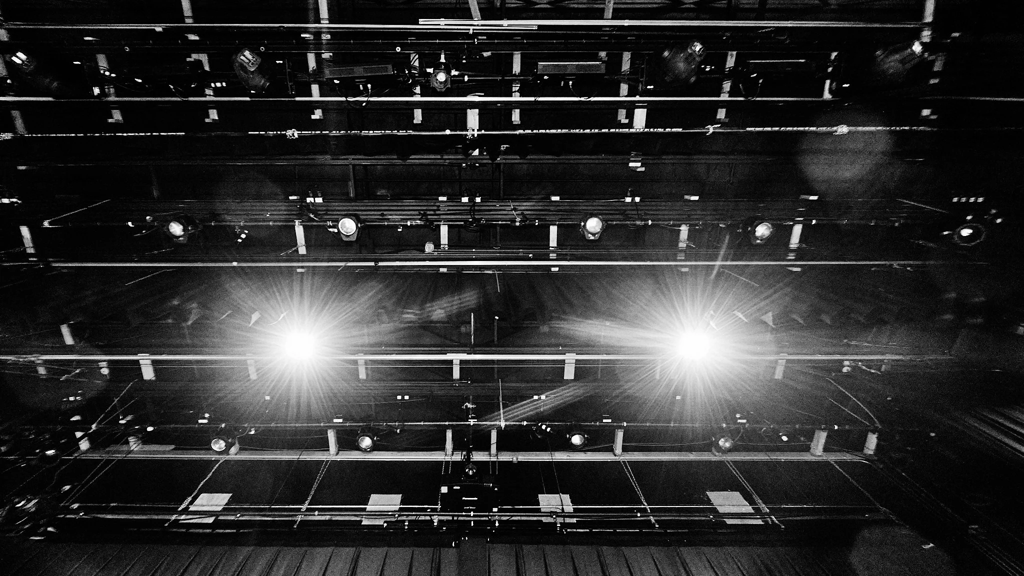 Black and white photograph of a theater stage lighting setup showing multiple stage lights directed downward, with some lights brightly lit, against a dark background.