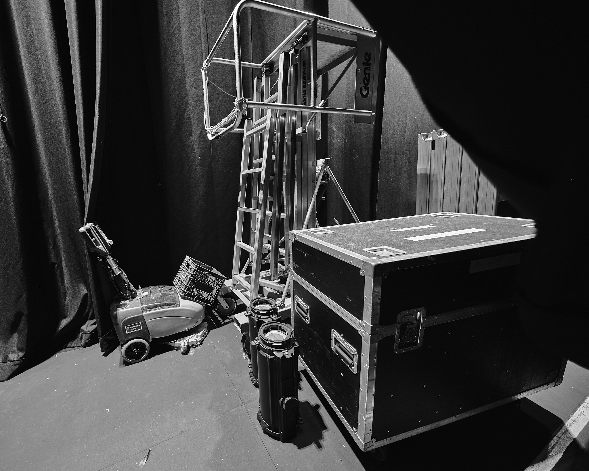Black and white photo of a stage setup with equipment cases, a ladder, and various tools on the floor, against a dark backdrop.