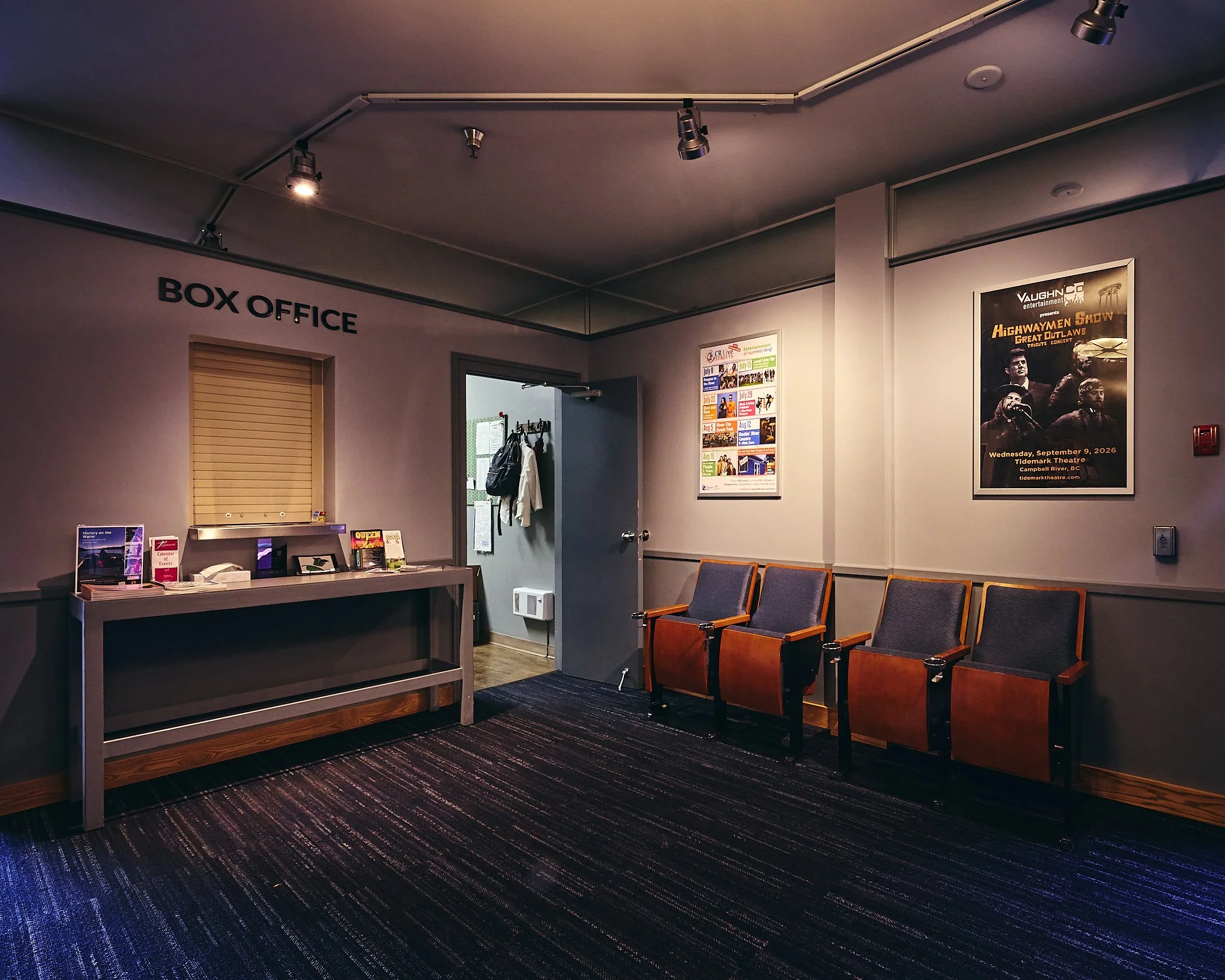 Inside a theater or auditorium, empty gates, chairs, and posters on the wall, with a dark carpeted floor and gray walls.