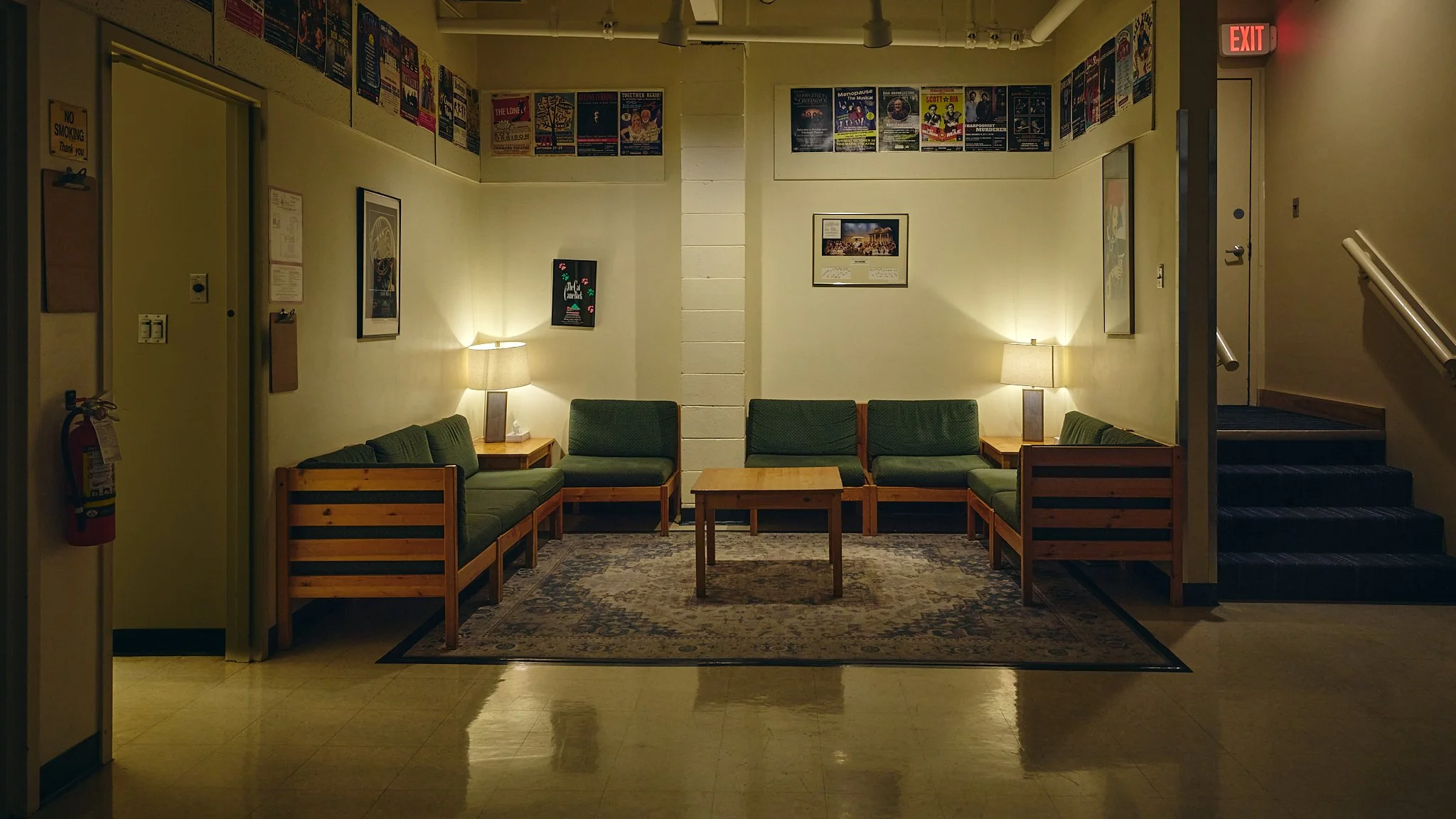 A cozy waiting area with green cushioned wooden chairs arranged around a small wooden table, lit by two table lamps on either side, with framed posters on the light yellow walls and a patterned rug on the floor.