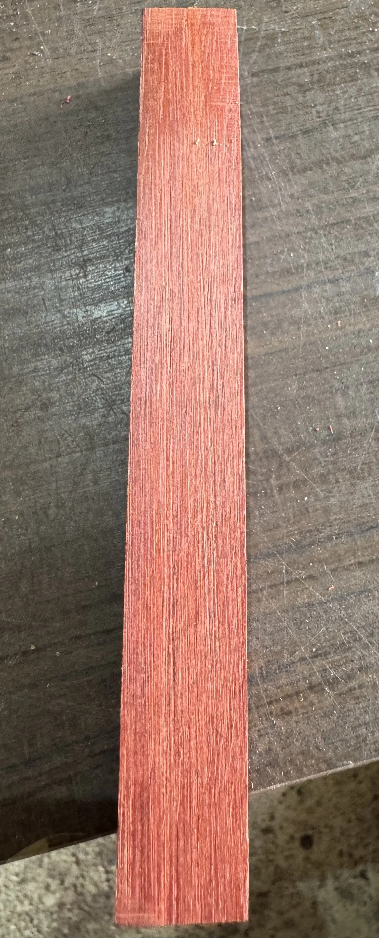 A close-up photograph of a rectangular piece of red wood with visible grain, laid on a dark, textured surface.