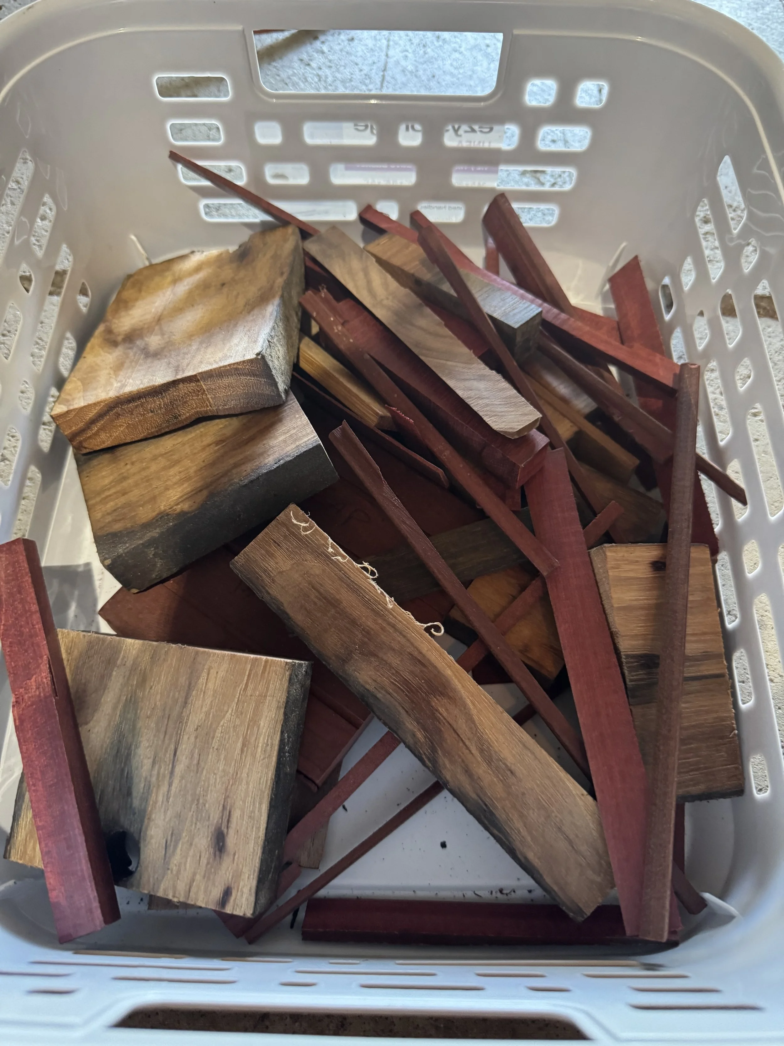 A white plastic laundry basket filled with various pieces of wood, including light and dark-colored wood, some with reddish hues, on a tiled floor.