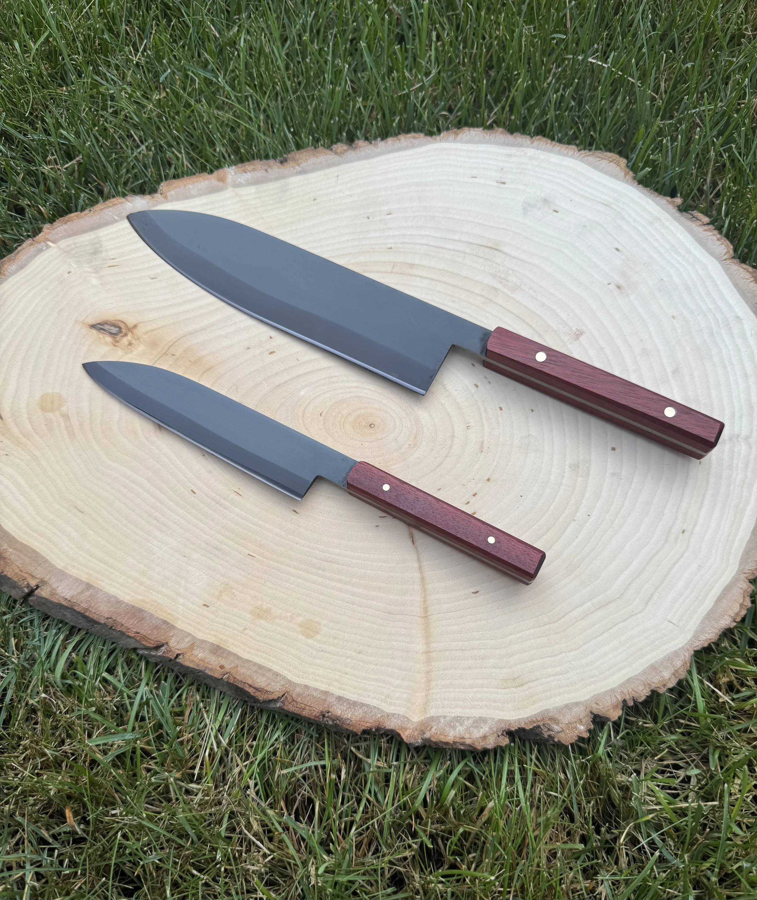 Two stainless steel kitchen knives with wooden handles resting on a flat cross-section of a tree trunk, outdoors on grass.
