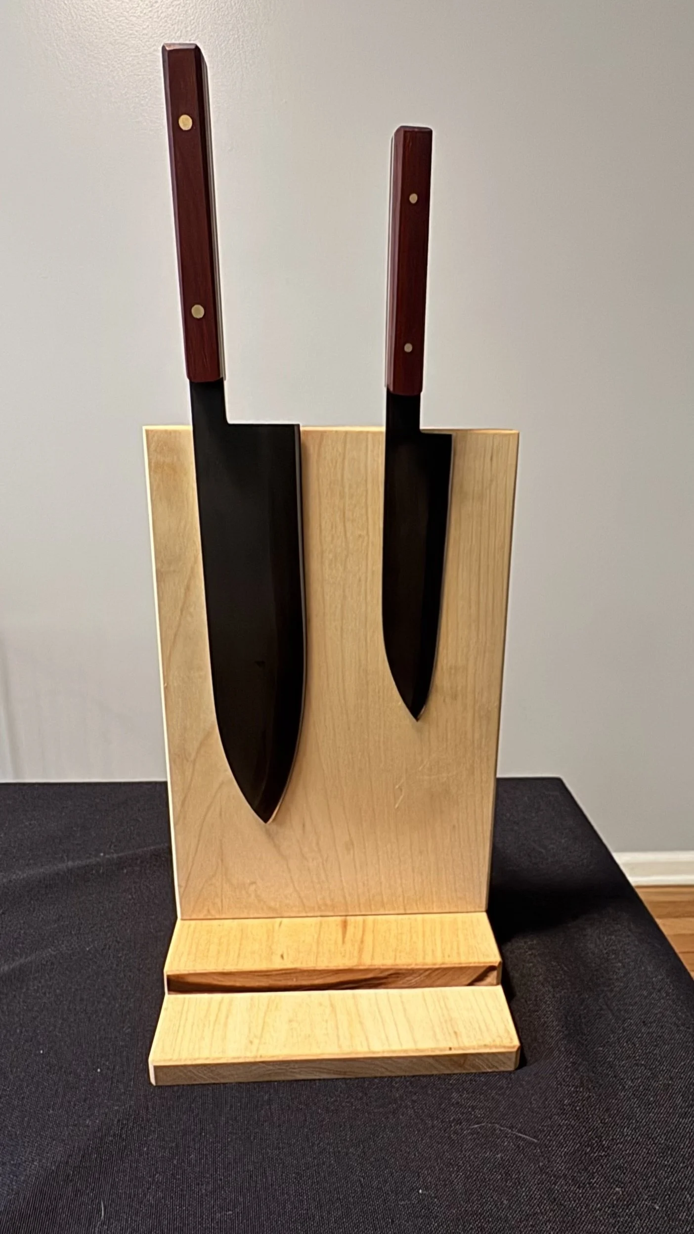 Two large chef knives with dark blades and wooden handles displayed on a wooden stand against a plain wall.