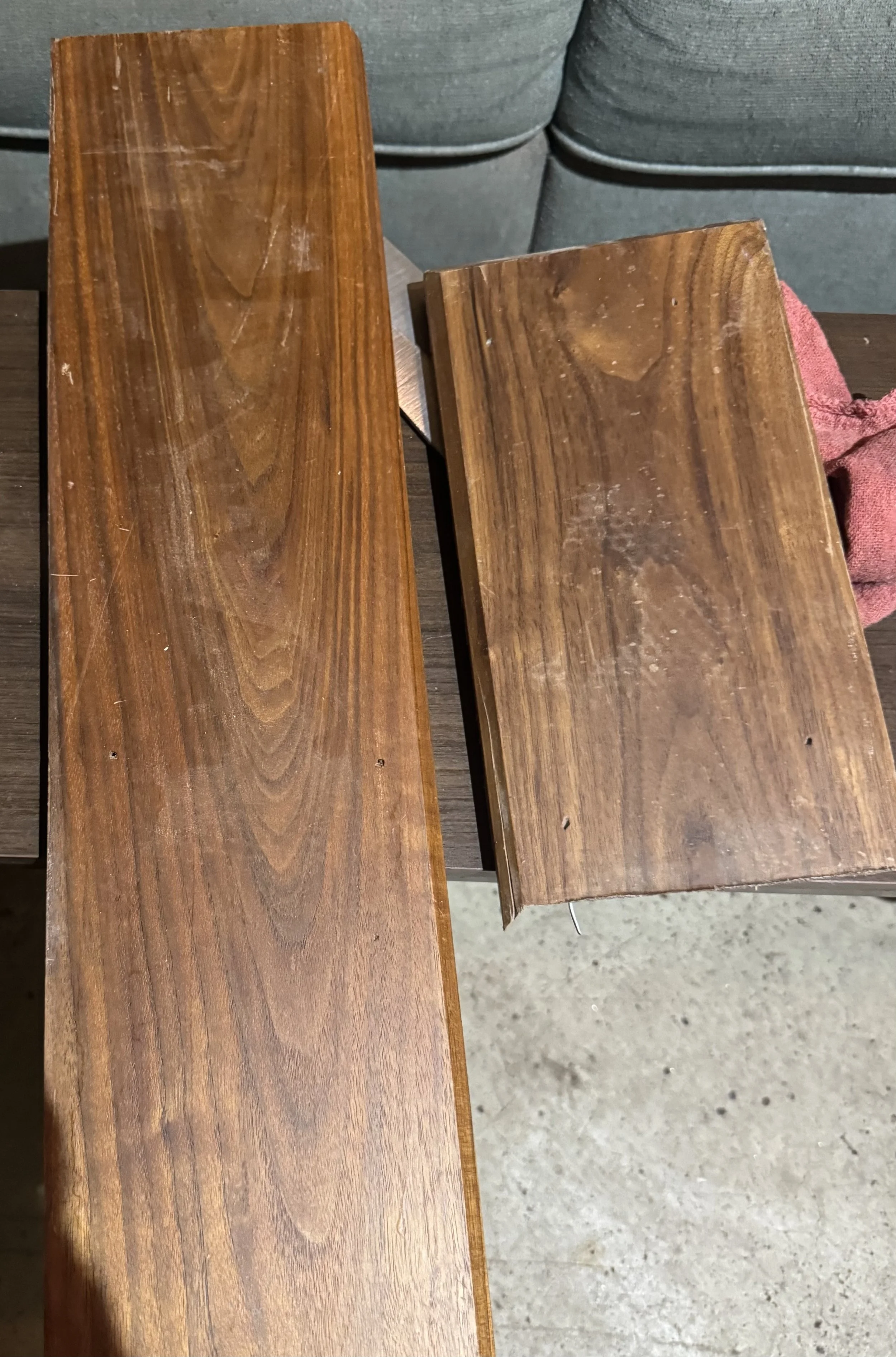 Two wooden planks of different sizes and colors resting on a table in front of a gray fabric sofa. One plank is darker and smoother, the other is lighter with visible grain patterns.