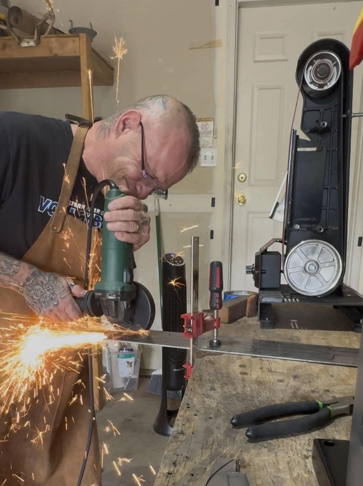 A man wearing glasses and a brown apron is welding a metal piece, causing sparks to fly. He is in a workshop with tools and equipment visible, including a large black and silver device on the right.