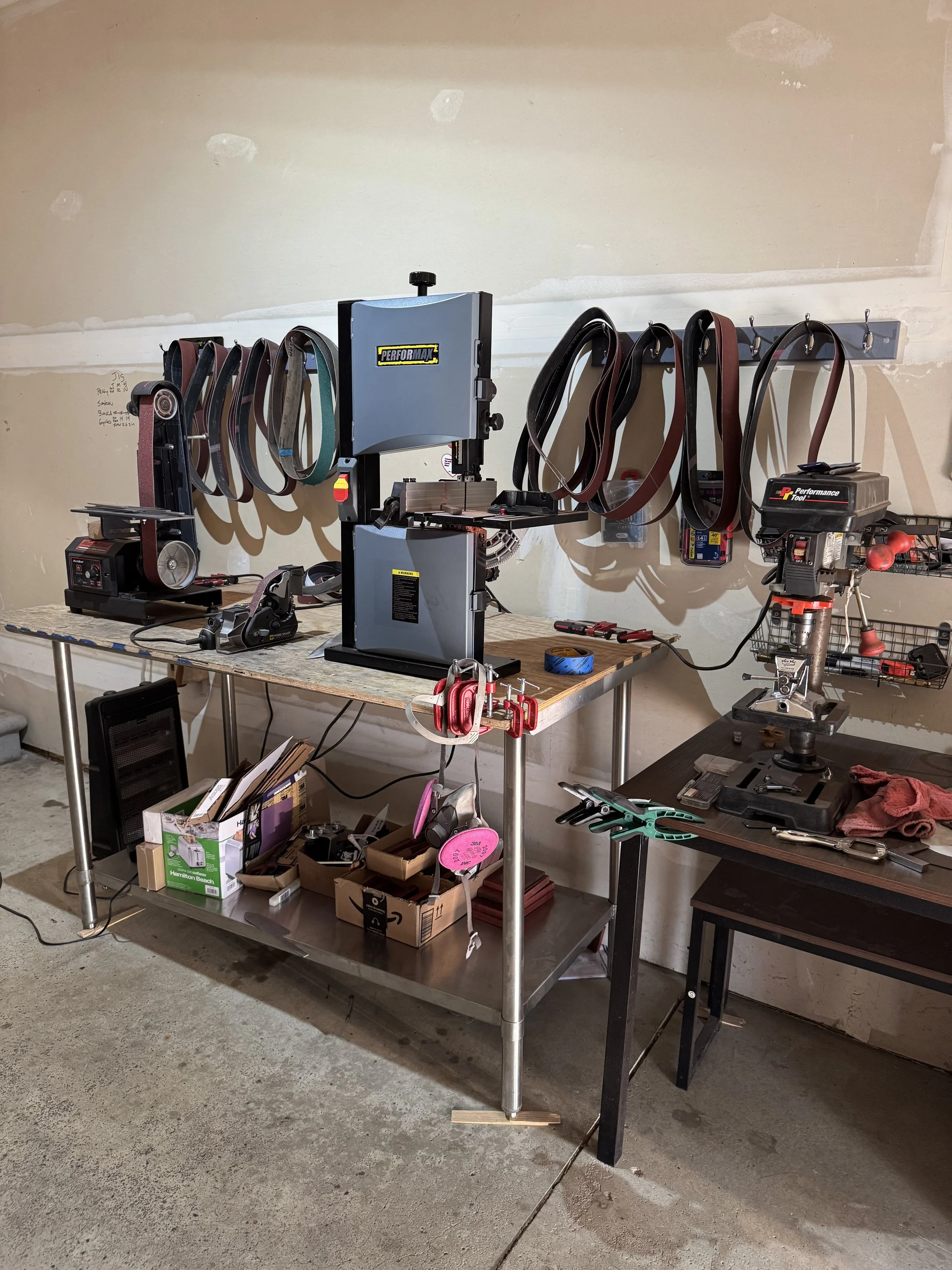 A workbench with various tools and equipment, including a bandsaw, belts, clamps, a drill press, and boxes of supplies, in a workshop with unfinished walls.