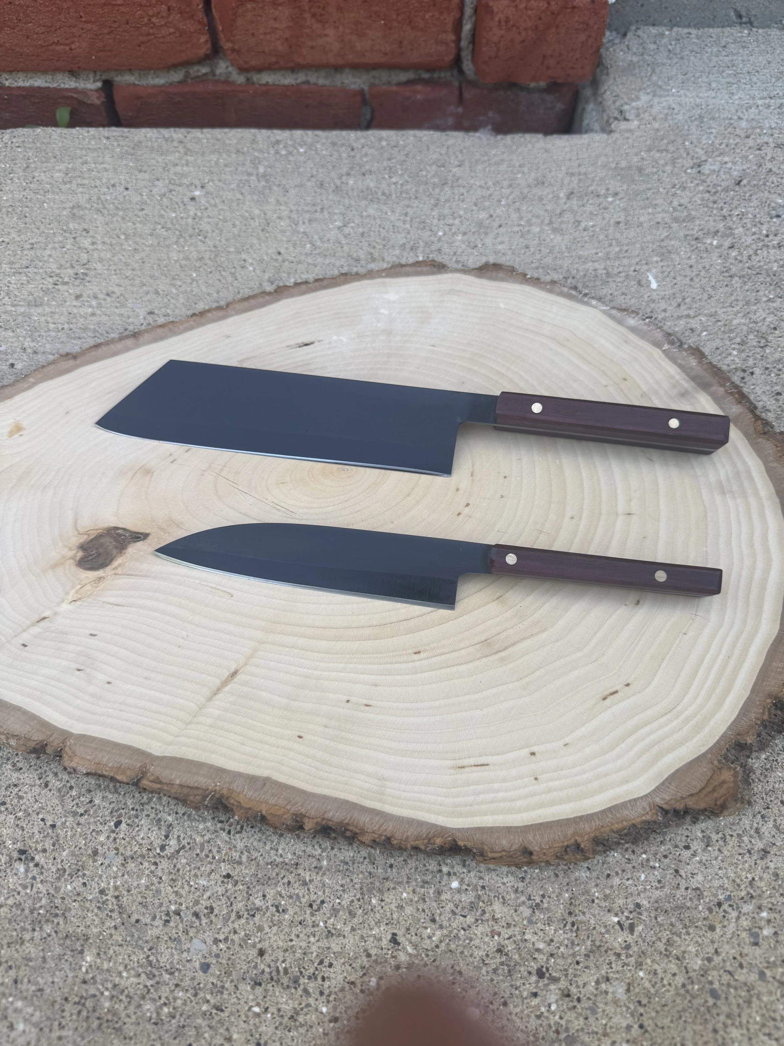 Two kitchen knives with dark blades and wooden handles resting on a large wooden cutting board with tree ring patterns, placed outdoors on concrete next to a brick wall.