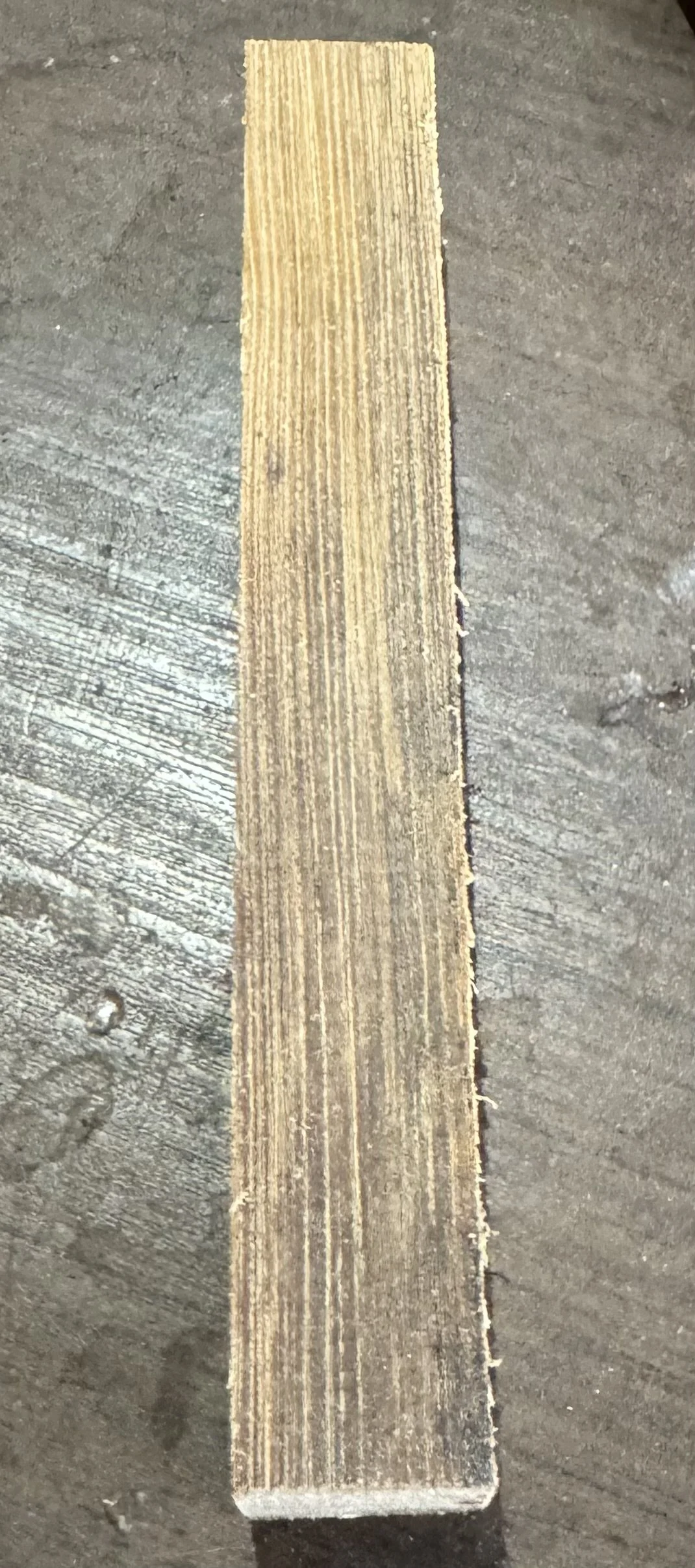 A piece of wood with visible grain, lying flat on a dark, textured surface.