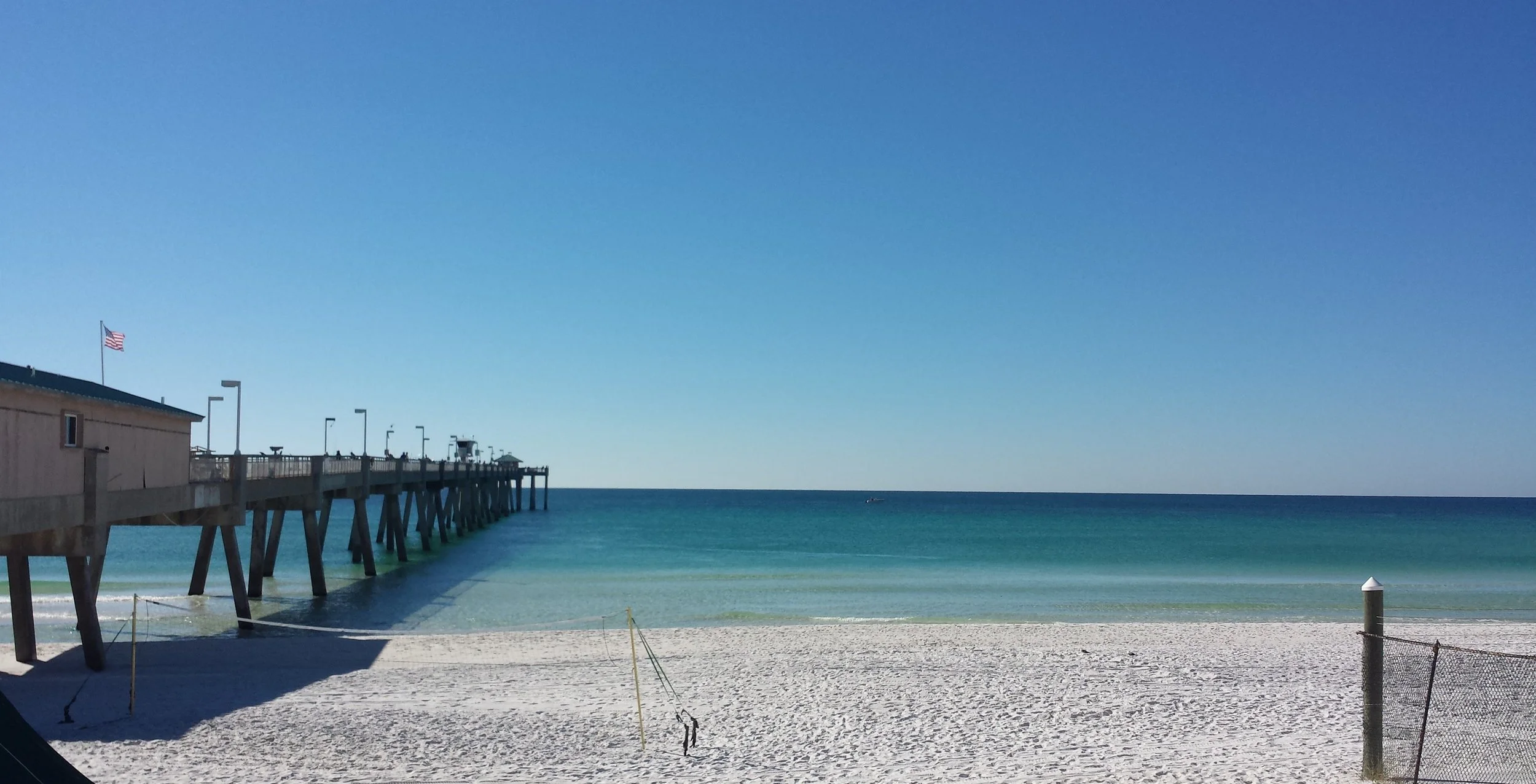 View of a Florida beach with a pier extending into turquoise water, clear blue sky, and a small building on the left with an American flag flying.