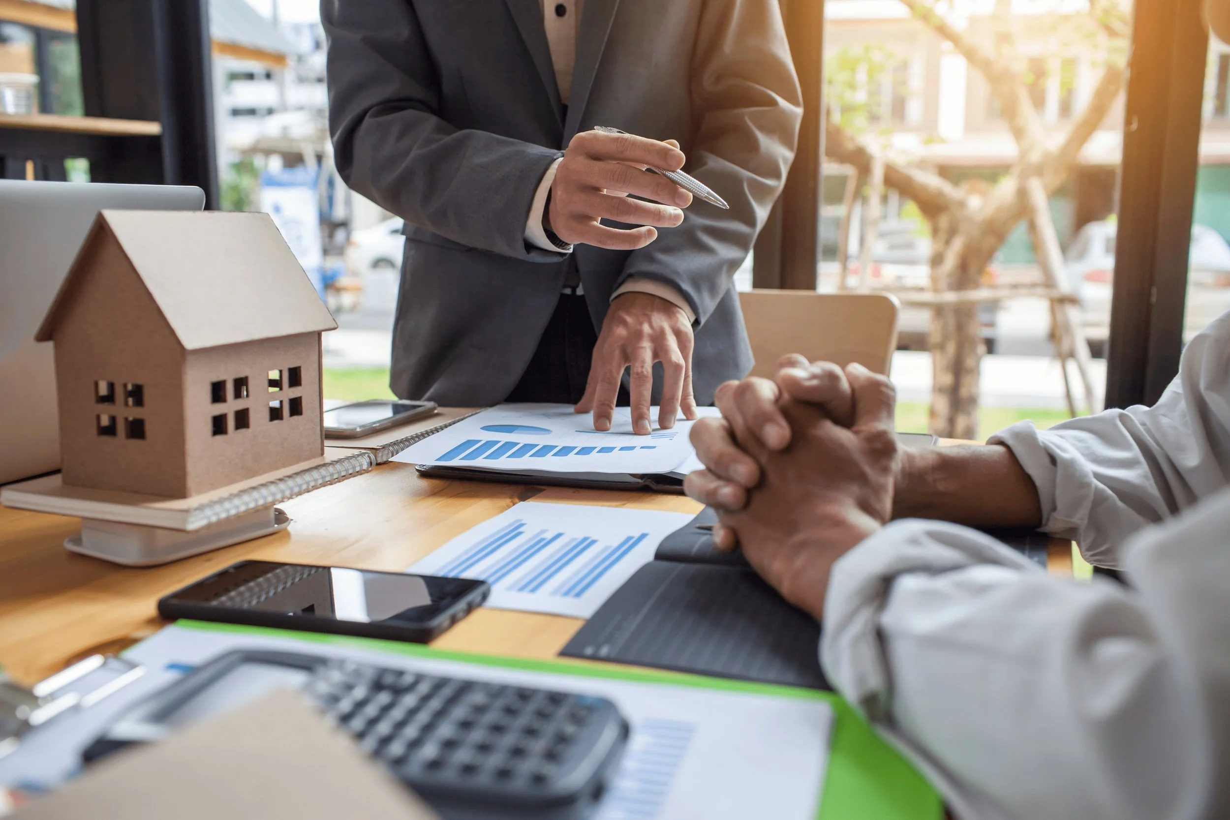 A person in a suit appears to be discussing documents with another person, with a small cardboard house model on the table, and other office items like papers, a calculator, and a laptop visible.