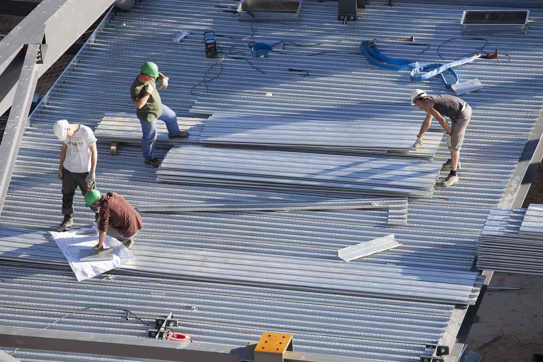 Construction workers installing or repairing a metal roof, some using tools and a blueprint, on a building.