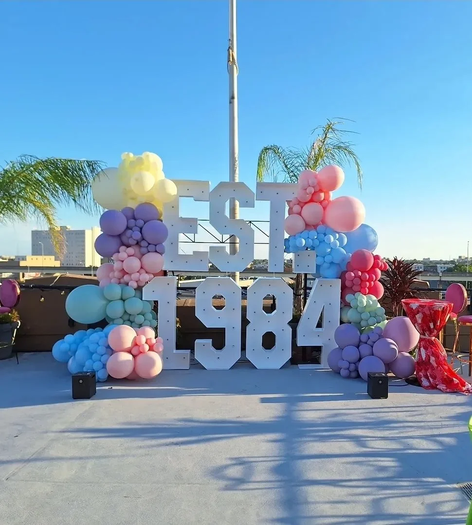 Large white letters showing 'EST 1984' decorated with pastel balloons in pink, purple, blue, and yellow surrounding the display, with a clear sky and cityscape in the background.