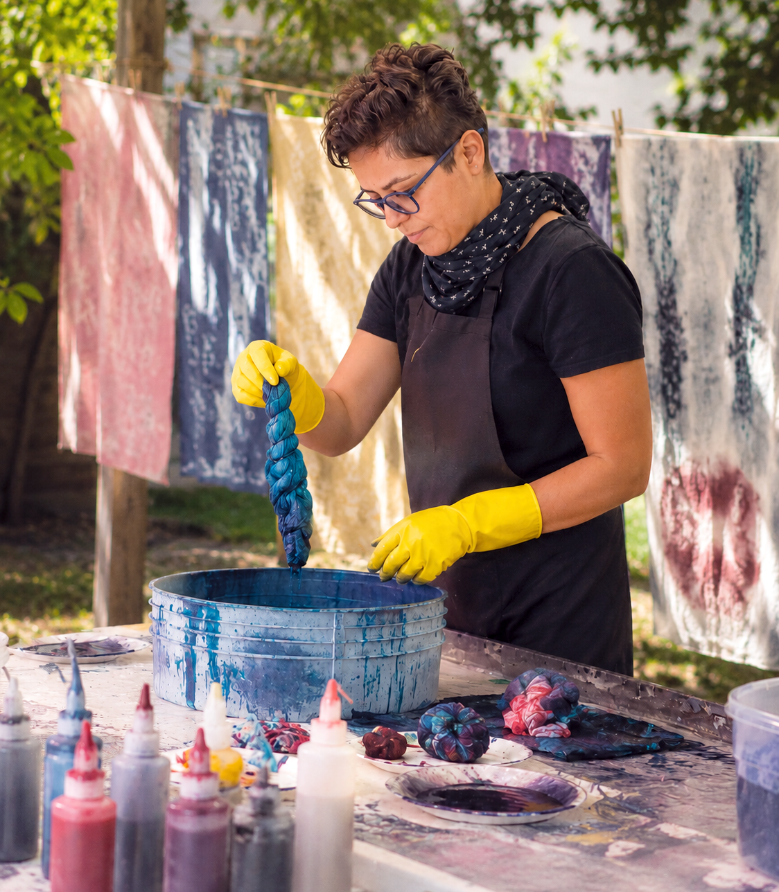 Artist Elnaz wearing yellow gloves, a black apron, glasses, and a bandana is dipping a twisted fabric into blue dye in a large container. Behind her, colorful dyed fabrics hang on a clothesline. Art supplies and more dyed fabrics are on the table.